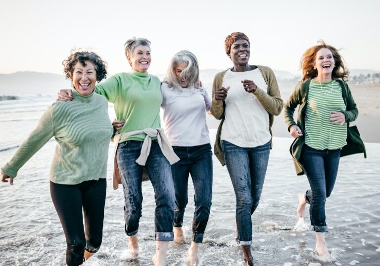 Group of middle-aged women laughing and enjoying a walk on the beach, representing diverse experiences and potential benefits of hormone replacement therapy for well-being and vitality